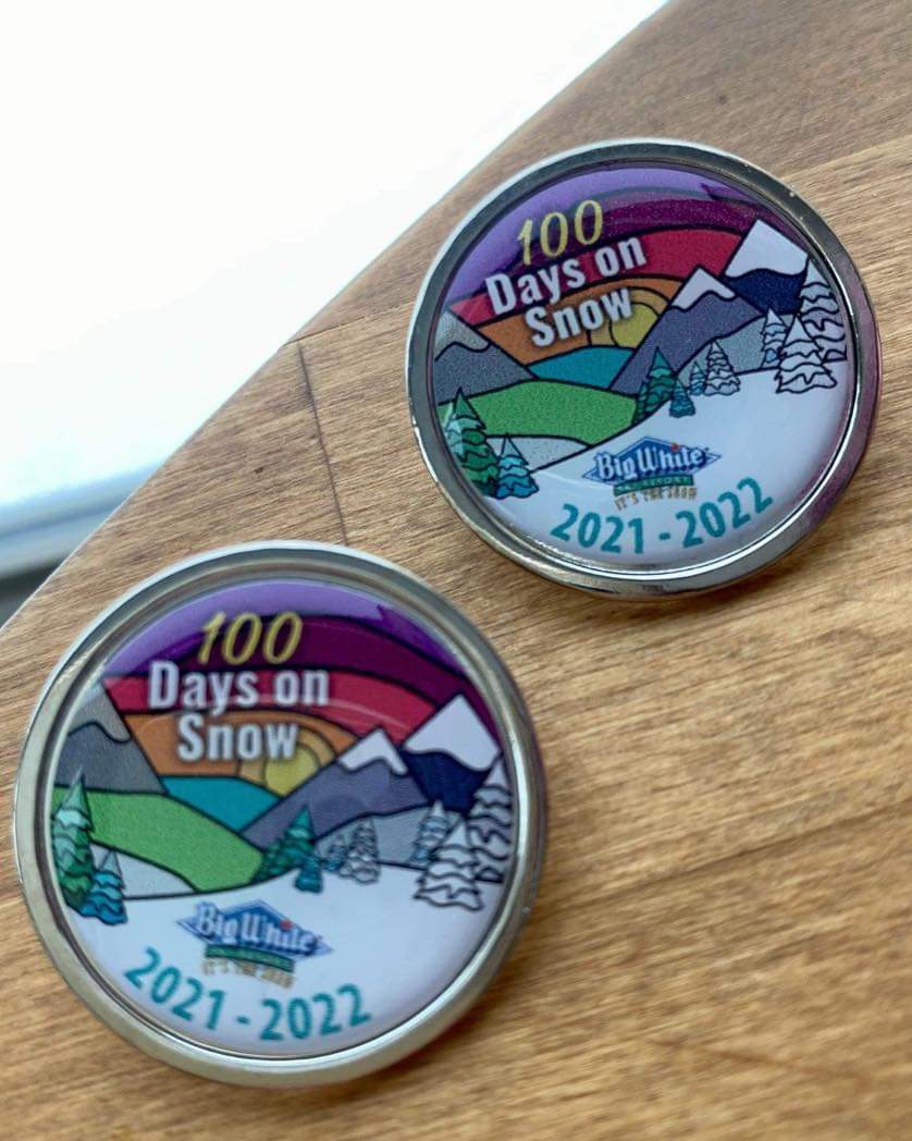 Two 100 Days on Snow Badges for the 2021 to 2022 season at Big White with colourful art sit on a wooden table.