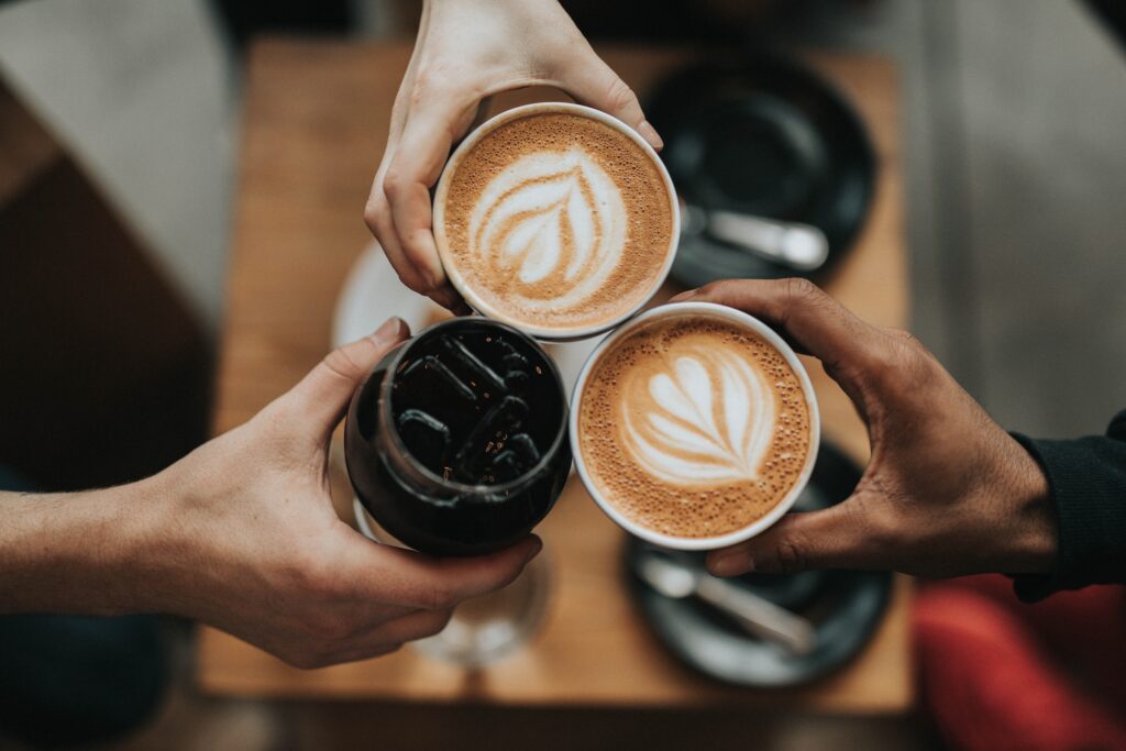 An overhead view of three people 'cheers'ing their coffee drinks with two lattes and an iced coffee over a table.